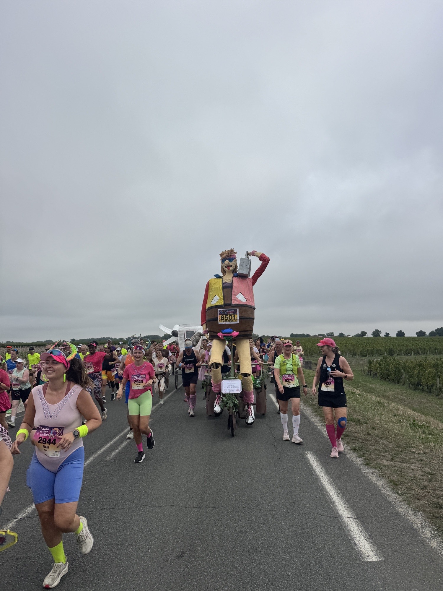 Coureurs déguisés et char géant de Château Lynch-Bages sur une route bordée de vignes du Médoc