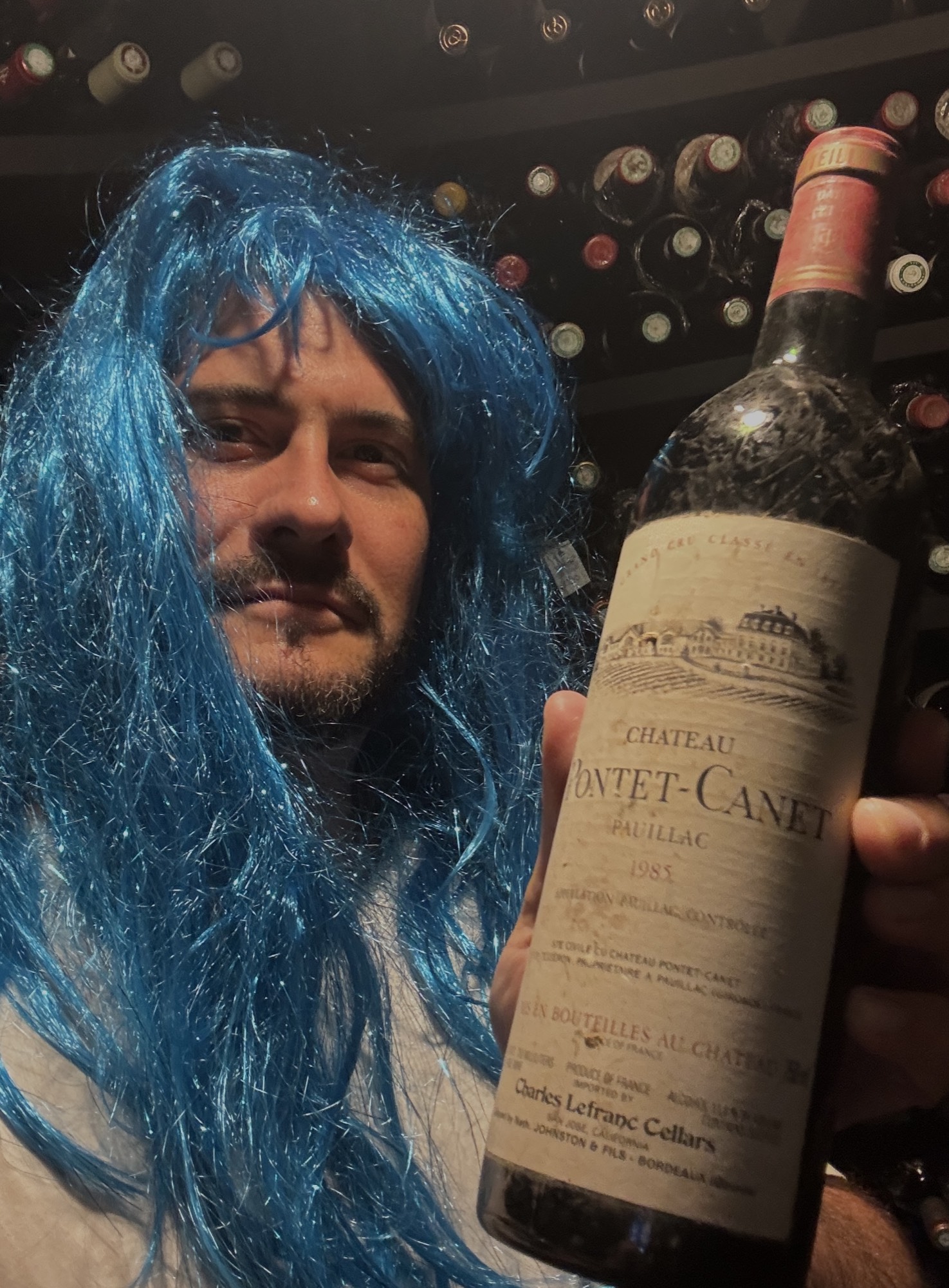Xavier Lacombe en perruque bleue tenant une bouteille de Château Pontet-Canet devant des casiers à bouteilles